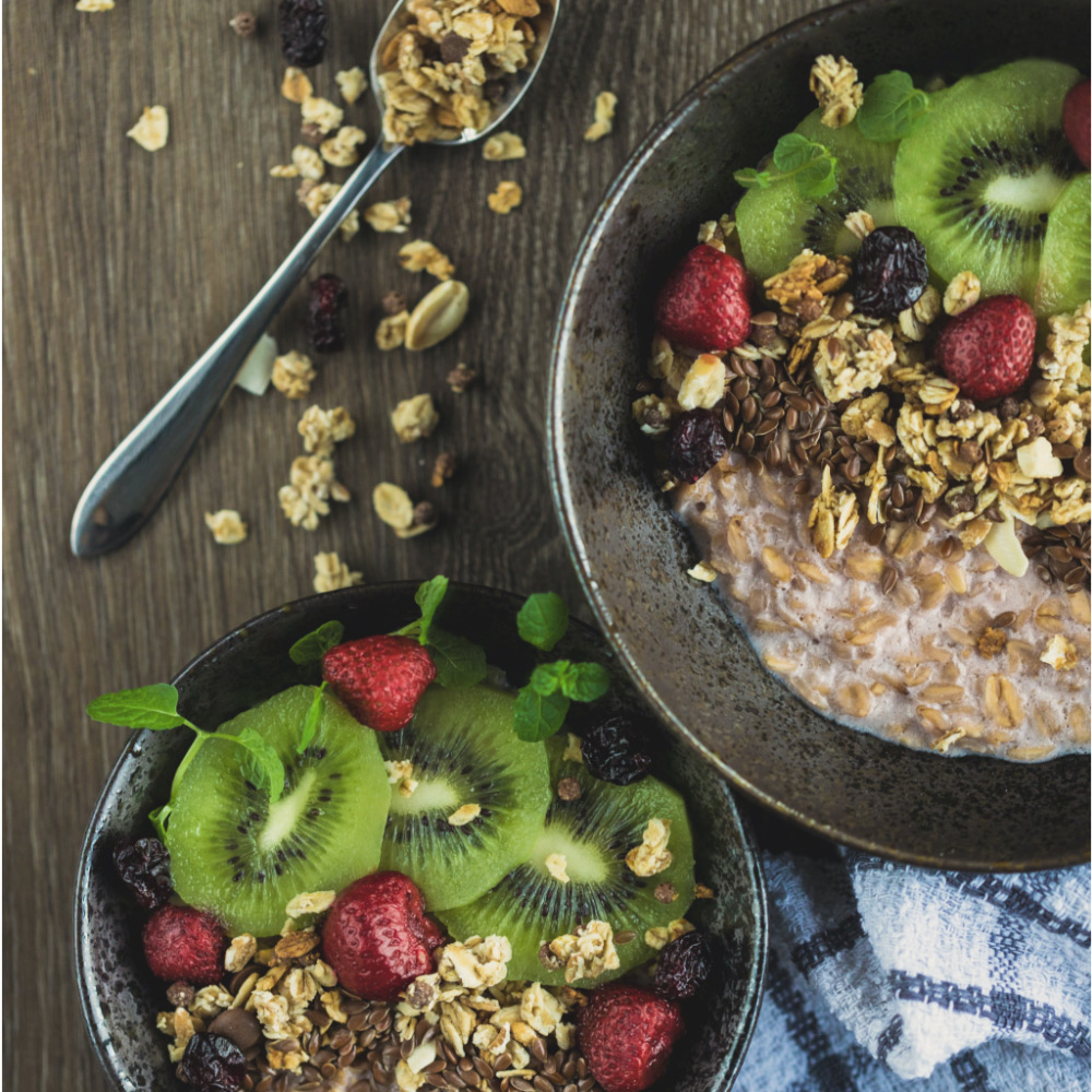 An image of two bowls of granola with fresh and dried fruits including kiwi and raspberries, plus seeds